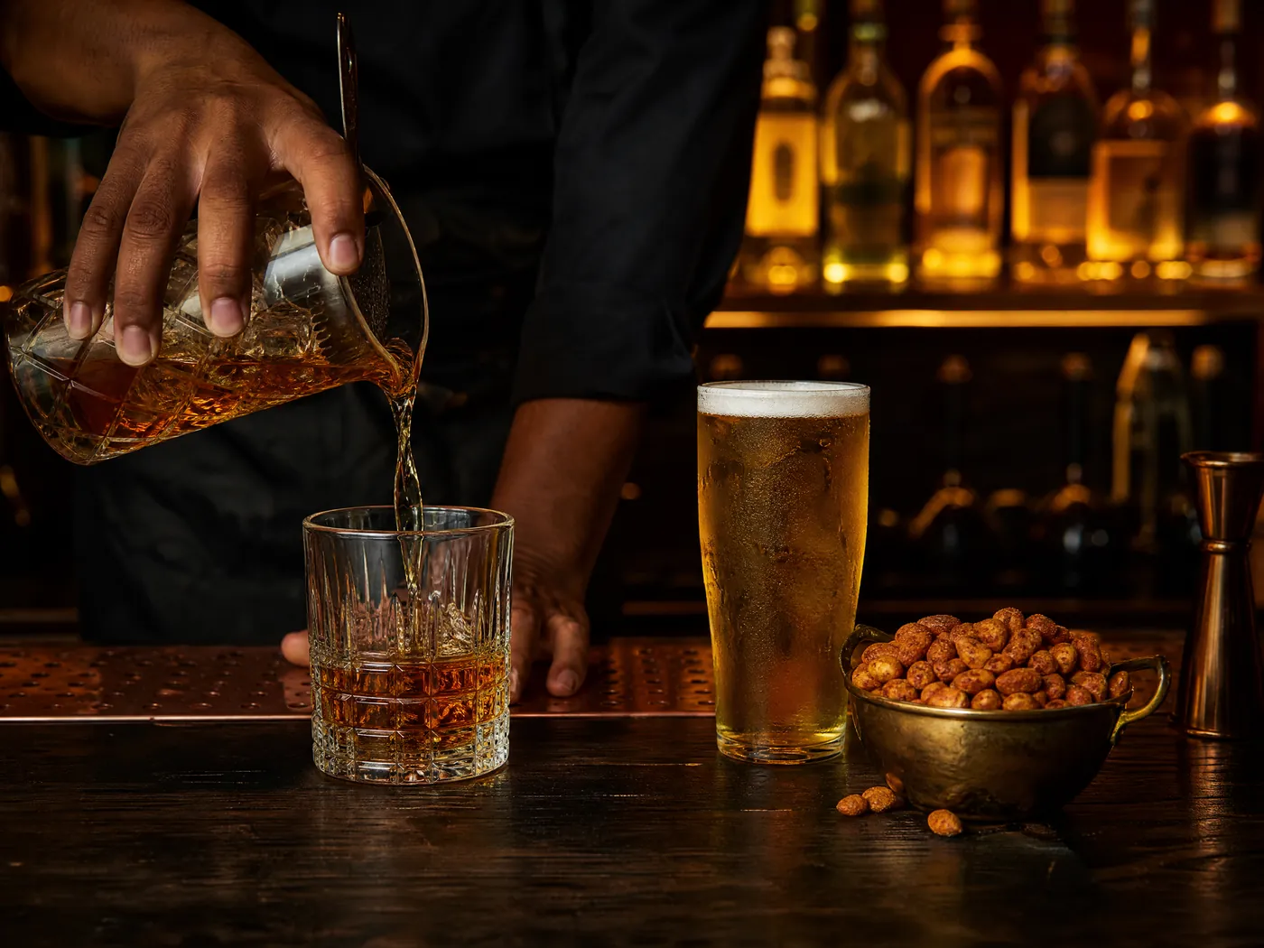A bartender pours amber whisky into a cut-glass tumbler beside a pint of lager and a bowl of masala peanuts.