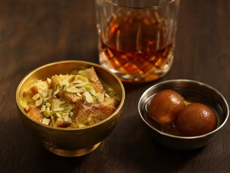 Double ka meetha topped with almonds and pistachios in a brass bowl, with gulab jamun beside it.